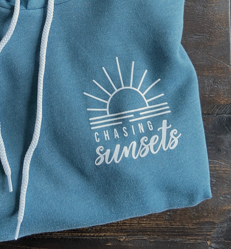 Puede incluir: Una sudadera con capucha azul con un gr&aacute;fico blanco de una puesta de sol y el texto "CHASING sunsets".