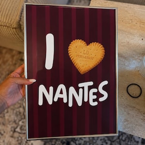 Peut inclure: Une affiche rectangulaire encadrée avec un fond à rayures verticales bordeaux foncé et noir. Le texte affiche I, suivi d'un biscuit LU Petit-Beurre en forme de cœur et du mot NANTES en grandes lettres blanches.