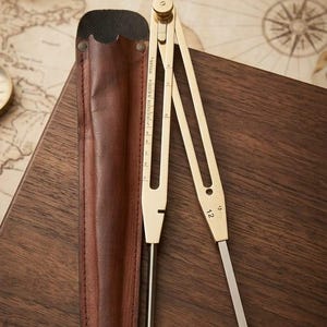 Puede incluir: Un compás de dibujo de latón con una regla integrada y el número 12 grabado en una pata, acompañado de una funda de cuero marrón. El compás cuenta con dos puntas de metal y una perilla de ajuste circular en la bisagra superior.