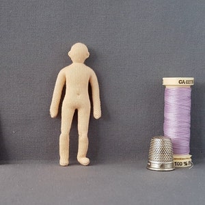 May include: A beige cloth doll with a simple design, standing on a grey surface. The doll has a head, arms, and legs, and is made of fabric. There is a spool of lavender thread and a silver thimble in the background.