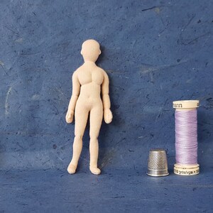 May include: A nude-coloured fabric doll with a simple design, standing on a blue surface. A spool of lavender thread and a silver thimble are on the right side of the image. A business card for Prairie Crocus Studio is on the left side of the image.