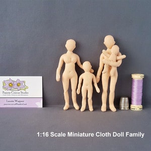 May include: A 1:16 scale miniature cloth doll family. The family includes a mother, father, and child. The dolls are unpainted and made of beige fabric. The dolls are standing on a grey surface. The image also includes a reel of purple thread and a thimble.