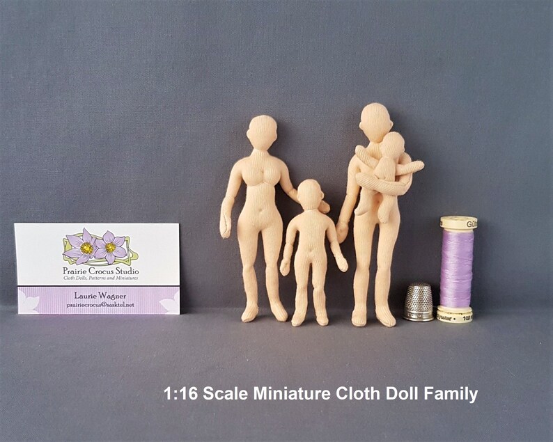 May include: A 1:16 scale miniature cloth doll family. The family includes a mother, father, and child. The dolls are unpainted and made of beige fabric. The dolls are standing on a grey surface. The image also includes a reel of purple thread and a thimble.