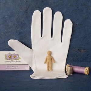 May include: A small, unpainted wooden doll figure stands on a white cloth glove. The doll is about 4 inches tall and has a simple, human-like form. A business card for Prairie Crocus Studio is visible in the lower left corner of the image. A purple spool of thread and a silver thimble are in the lower right corner.