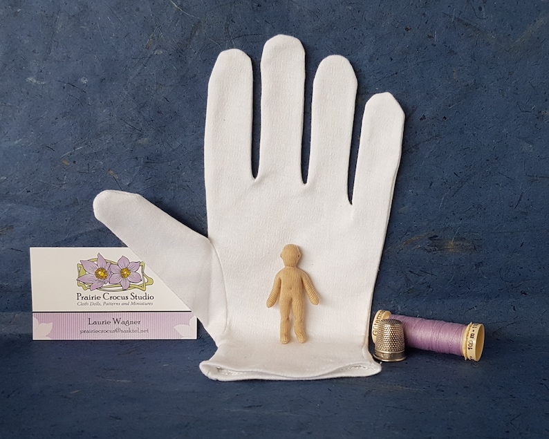 May include: A small, unpainted wooden doll figure stands on a white cloth glove. The doll is about 4 inches tall and has a simple, human-like form. A business card for Prairie Crocus Studio is visible in the lower left corner of the image. A purple spool of thread and a silver thimble are in the lower right corner.