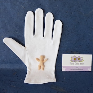 May include: A white cotton glove with a small, nude-coloured doll figure in the palm. The glove is lying on a blue surface. A business card with the text "Prairie Crocus Studio" is to the right of the glove.