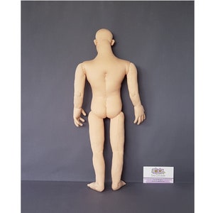 May include: A beige fabric doll body, viewed from the back, with arms and legs extended. The doll has a bald head and is standing against a gray background. The doll is made of a soft, light-colored fabric. A small business card is in the lower right corner.