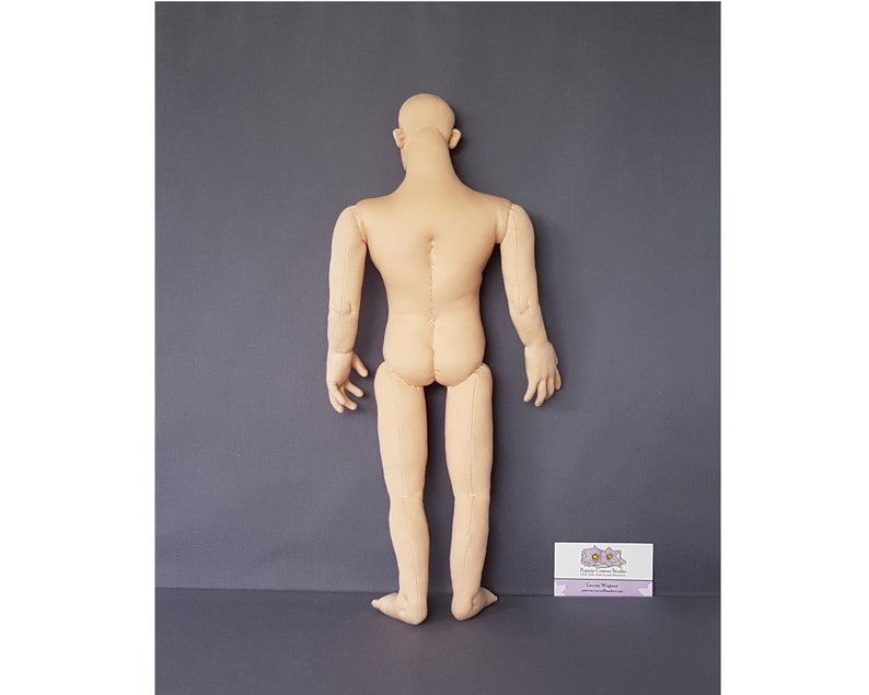 May include: A beige fabric doll body, viewed from the back, with arms and legs extended. The doll has a bald head and is standing against a gray background. The doll is made of a soft, light-colored fabric. A small business card is in the lower right corner.