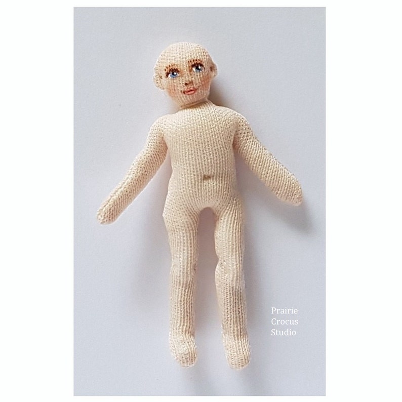 May include: A handmade knitted doll with a painted face, blue eyes, and a light pink complexion. The doll is standing with its arms outstretched.