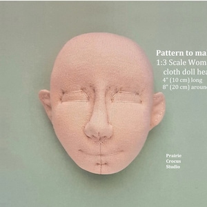 May include: A pink cloth doll head with stitched facial features. The head is 4 inches (10 cm) long and 8 inches (20 cm) around. The text "Pattern to make 1:3 Scale Woman's cloth doll head 4" (10 cm) long 8" (20 cm) around Prairie Crocus Studio" is visible in the image.