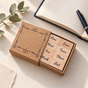 May include: A set of eight rectangular wooden stamps housed in a brown cardboard box with a decorative black border. The stamps feature the printed text Mon, Tue, Wed, Thur, Fri, Sat, Sun, and Week in a black cursive script font.