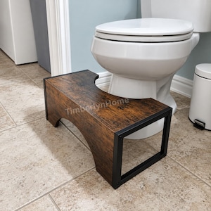 May include: A bathroom toilet stool with a dark brown wood grain finish and a black metal frame. The stool features a curved cutout design to fit around the base of a toilet. The text TimberlynHome is printed on the top surface.