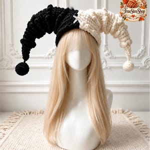 May include: A two-toned jester hat made of chunky black and white yarn. The hat features two long, curved, pointed sections with a pom-pom at each end. A small white ribbon bow is centred at the front where the two colours meet.