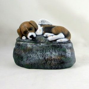 May include: A ceramic pet urn shaped like a rock with a sleeping beagle with wings on top. The urn is painted in shades of green, brown, and white. The name "Summer" is inscribed on the urn.