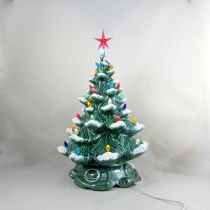 May include: A ceramic Christmas tree in a deep green colour, adorned with colourful lights and a red star on top. The tree has a snow-like effect on the branches and sits on a decorative base. A white power cord extends from the base.