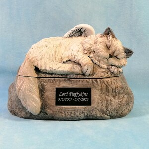 May include: White ceramic pet urn with a sleeping cat with angel wings on top. The urn is shaped like a rock and has a plaque on the front that reads "Lord Fluffykins 9/6/2007 - 5/7/2023".