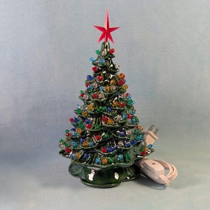 May include: A glossy dark green ceramic Christmas tree ornament featuring a pointed red star topper and numerous small, multi-coloured translucent plastic bulbs embedded throughout the branches, accompanied by a white electrical cord and plug.