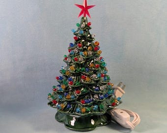 May include: A glossy dark green ceramic Christmas tree decoration featuring a pointed red star topper and numerous small, multi-colored translucent plastic bulbs embedded throughout the branches, accompanied by a white electrical cord and plug.