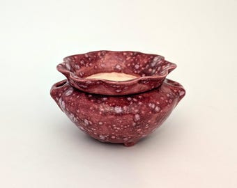 Può includere: Un set di due ciotole in ceramica. La ciotola inferiore, più grande, è di un bordeaux intenso con macchie bianche e due manici. La ciotola superiore, più piccola, ha un bordo smerlato e una finitura maculata bordeaux simile. Il set è decorativo.