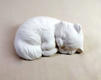May include: White ceramic sculpture of a sleeping cat curled up in a ball. The cat has a detailed face and fur texture.
