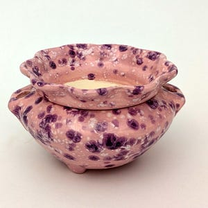 May include: A two-piece ceramic container with a pink base and a matching lid. The container and lid are decorated with dark purple spots. The lid has a scalloped edge. The interior of the container is white.