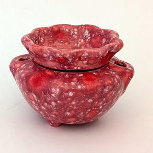 Peut inclure: Un brûleur d'huile en céramique rouge en deux parties avec une finition texturée et mouchetée. La base a une forme arrondie avec trois petits pieds et deux poignées latérales. La partie supérieure est un petit bol avec un bord festonné.