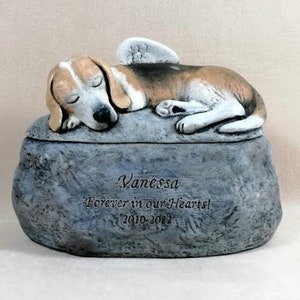 May include: A ceramic pet urn shaped like a rock with a sleeping beagle dog with angel wings on top. The urn is inscribed with the name "Vanessa", the text "Forever in our hearts!" and the dates "2010-2022".