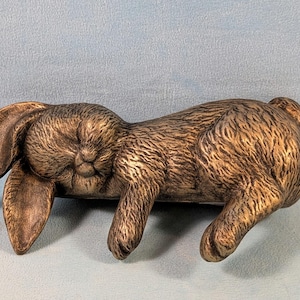 May include: A brown rabbit ornament with detailed fur texture and long ears. The rabbit is lying down, with its head and ears to the left and its rear to the right. The ornament is made of a solid material.