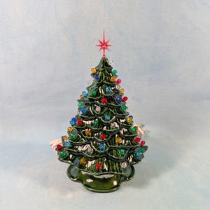 May include: A ceramic Christmas tree with a glossy green finish. The tree is decorated with colourful lights and a red star on top. The base of the tree is also green and has a scalloped edge. A white electrical cord is attached.