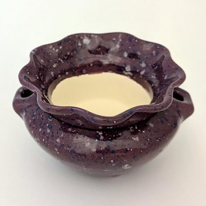 May include: A small, round ceramic bowl with a dark purple, speckled glaze and a ruffled, wavy rim. The interior of the bowl features a smooth, cream-coloured finish, contrasting with the textured, dark exterior. The vessel has a wide opening.