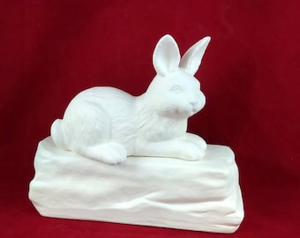 May include: A white ceramic rabbit figurine, resting on a rectangular base. The rabbit is in a relaxed pose, with its ears perked up. The sculpture is unpainted and has a smooth finish. The background is a solid red.
