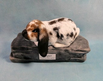 May include: A ceramic figurine of a black and white spotted rabbit with brown markings lying on a gray rock. The rabbit is named "Jasper" and the inscription reads "Loved & we meet again".