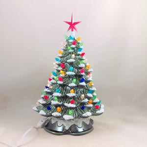 May include: A green ceramic Christmas tree with a red star on top and colorful lights. The tree has a white base with a snowy effect.