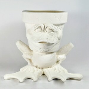 May include: A white ceramic planter with a whimsical duck-like face design. The planter features a wide opening and is supported by stylized duck feet. The face has detailed features, including closed eyes and a beak.