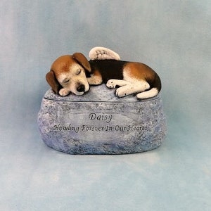 May include: A ceramic pet urn shaped like a rock with a sleeping beagle dog on top. The dog has a white angel wing and the urn is inscribed with the words "Daisy Howling Forever In Our Hearts".