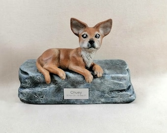 May include: A ceramic dog figurine, lying on a gray stone base. The dog is brown with a white chest and has a name tag that reads "Chuey" with dates. The figurine is a memorial or decorative item.