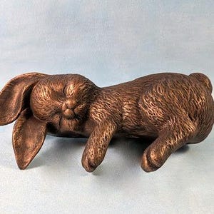 May include: Bronze rabbit figurine in a relaxed pose. The bunny has long, floppy ears and is lying on its side with its eyes closed. The sculpture has a textured surface and is set against a light blue background.