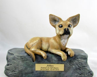 May include: A ceramic figurine of a tan chihuahua lying on a gray rock base. The base has a gold plaque with the inscription "Abbey Forever In Our Hearts 30/12/2010 - 02/07/2016".
