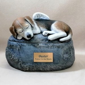 Könnte beinhalten: Eine Keramik-Tierurne in Form eines Steins mit einem schlafenden Beagle-Hund mit Engelsflügeln darauf. Die Urne hat eine kleine Plakette mit der Aufschrift "Buster Forever In Our Hearts".