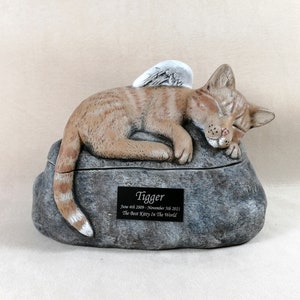 May include: A tan and brown cat figurine with angel wings, resting on a grey stone-like base. The base has a black plaque with the name "Tigger" and dates. A memorial item for a pet.