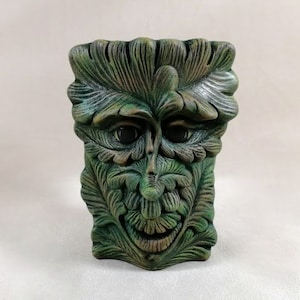 Puede incluir: Una escultura de rostro de hombre verde con una expresión sonriente. La escultura está hecha de un material verde oscuro con detalles de hojas y rasgos faciales. Los ojos son oscuros y el diseño general recuerda a la naturaleza.