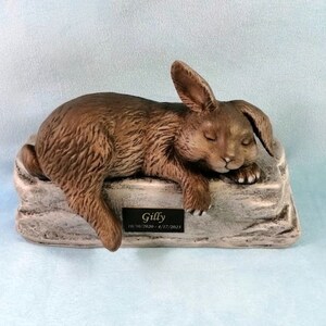May include: A ceramic figurine of a brown rabbit sleeping on a stone-like base. The base has a small plaque with the name "Gilly" and the dates "10/30/2019 - 4/17/2021".