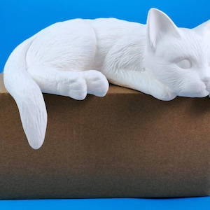 May include: White ceramic cat figurine lying on a brown cardboard box. The cat is in a sleeping position with its head resting on its paws.