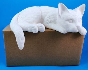 May include: White ceramic cat figurine lying on a brown cardboard box. The cat is in a sleeping position with its head resting on its paws.