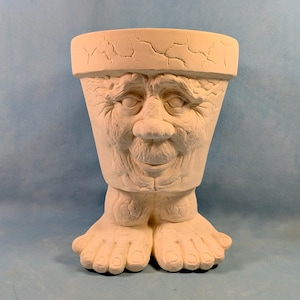 May include: A matte white ceramic planter shaped like a head with a cracked texture on the rim and face. The planter features a detailed human-like face with deep-set eyes, a prominent nose, and a mouth, supported by two large, textured feet at the base.