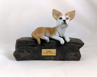 May include: A ceramic dog figurine, tan and white, reclines on a dark gray, stone-like base. The dog's name, TIMBIT, and dates 11-19-08 to 05-04-18 are on a gold plaque. The figurine is a memorial or decorative item.