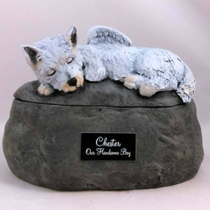 Pode incluir: Uma urna de cerâmica branca para animais de estimação em forma de rocha com um lobo branco com asas deitado em cima. A urna tem uma placa preta com o texto "Chester Our Handsome Boy".