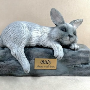 May include: A grey rabbit sculpture resting on a stone-like base. The rabbit is lying down with its eyes closed. A gold plaque on the base reads "Billy Always in our hearts."