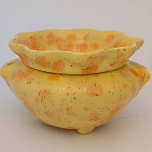 Peut inclure: Un cache-pot en céramique jaune avec un bord festonné et un motif orange tacheté. Le récipient en forme de bol présente deux petites poignées latérales et repose sur trois pieds courts, arborant une finition émaillée brillante et tachetée.