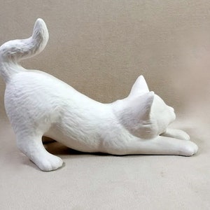 Op de afbeelding: Een wit keramiek kattenbeeldje in een speelse pose, met zijn staart om zijn lichaam gekruld en zijn hoofd naar de zijkant gedraaid. Het beeldje is ongeverfd en klaar om te worden gedecoreerd.